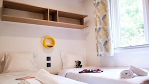A hotel room with two beds, a window with curtains, a wooden shelf, and a yellow circular object on the wall. - ZEcamping 4 étoiles piscine animaux acceptés mobil-home TV LV clim (Orgon)