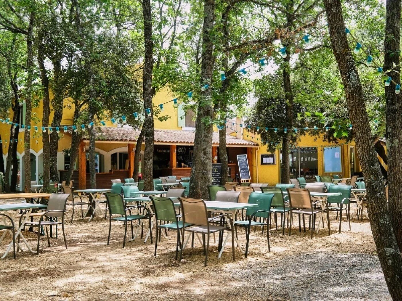 An outdoor cafe with tables and chairs, surrounded by trees and a yellow building.