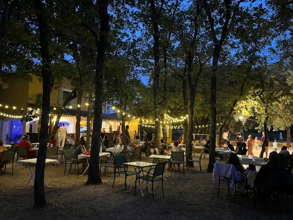 An outdoor dining area with tables and chairs, surrounded by trees and string lights.