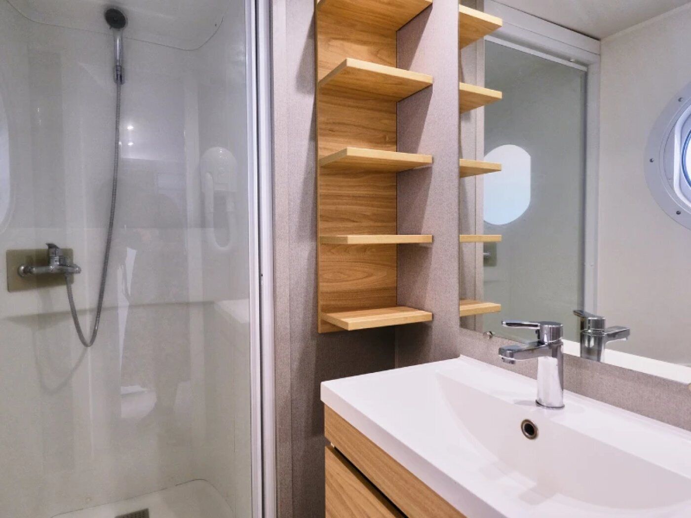 A modern bathroom with a glass shower enclosure, a white sink with a mirror, and wooden shelves.