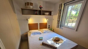 Un lit bien fait, avec un chapeau et un livre posés dessus, une table de chevet, une étagère avec des livres et un panier, et une fenêtre avec des rideaux.