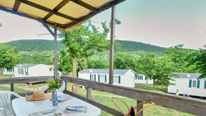 A covered outdoor seating area with a table and chairs, overlooking a row of mobile homes in a green, hilly landscape. - ZEcamping 3 étoiles familial piscine chauffée dog friendly cottage clim 30m2 (Lussas)