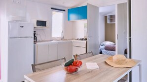 A compact kitchen with white cabinets, a microwave, and a refrigerator. A dining table with a basket and a bowl of fruit is in the foreground. - ZEcamping 3 étoiles familial piscine chauffée dog friendly cottage clim 30m2 (Lussas)