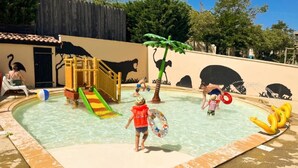 A children's water play area with slides and a pool, surrounded by a fence and trees.