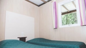 A room with two beds, a small table, a window with curtains, and a view of greenery outside.