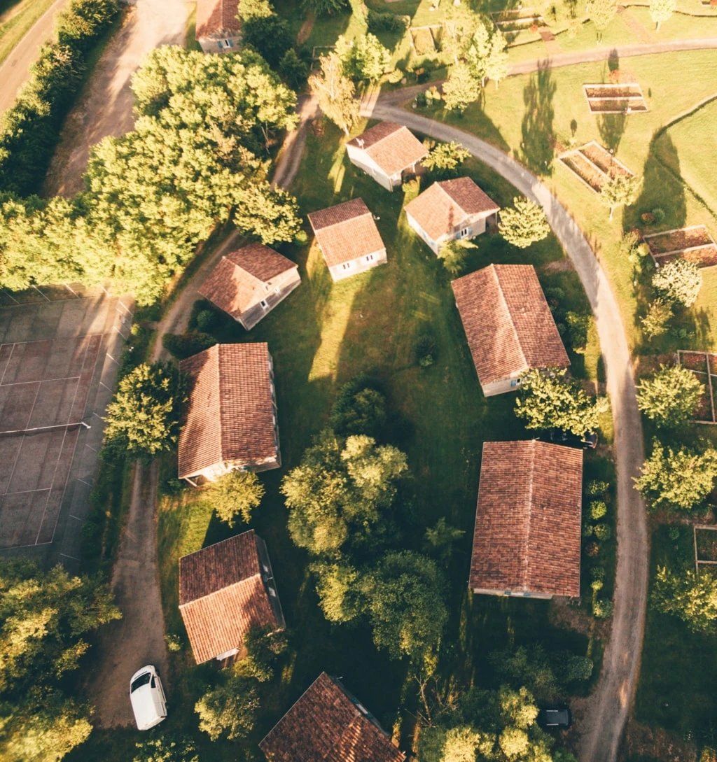 An aerial view of a residential area with houses arranged in a circular pattern, surrounded by trees and a clear sky.
