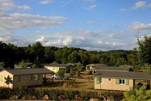 A row of single-story cabins with a flat roof, surrounded by greenery and trees, under a partly cloudy sky. - ZEcamping 3 étoiles animaux acceptés tennis piscine couverte (Beynat)