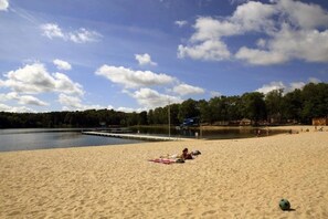 Een zandstrand met een kalm meer, een steiger en bebossing in de verte.