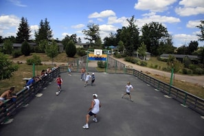A sports court with a basketball hoop, surrounded by a fence, trees, and houses in the background.