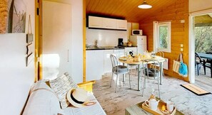 A cozy cabin interior with a dining area, kitchen, and a sofa with a hat and sunglasses. - Chalet 3 étoiles - Piscine  - eebbhh (Saint-Denis-d'Oléron)