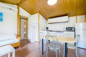 Une cuisine compacte avec des meubles blancs, un four à micro-ondes et un réfrigérateur. On y trouve une table à manger avec des chaises et un banc avec un coussin.