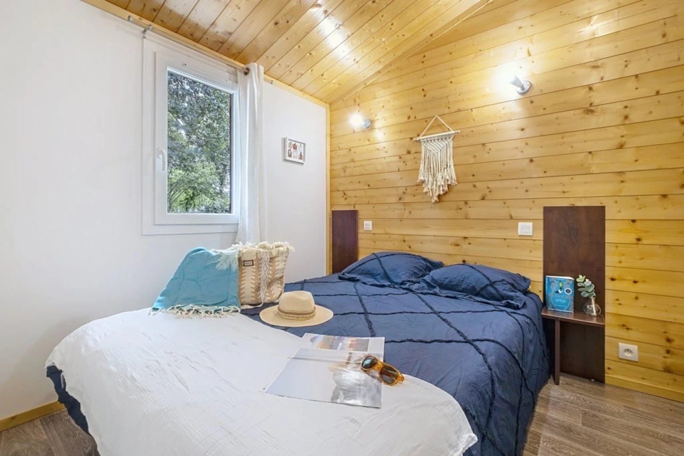 Une pièce avec un plafond en bois, un lit recouvert d’une couverture bleue, un oreiller blanc, un chapeau et une petite table sur laquelle se trouvent un livre et une plante.
