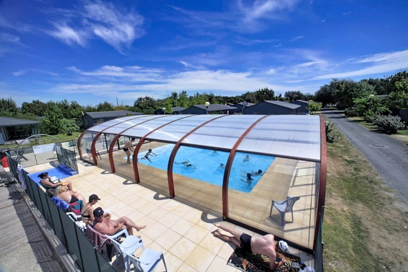 Una piscina techada con techo transparente, rodeada por una terraza de madera y sillas.