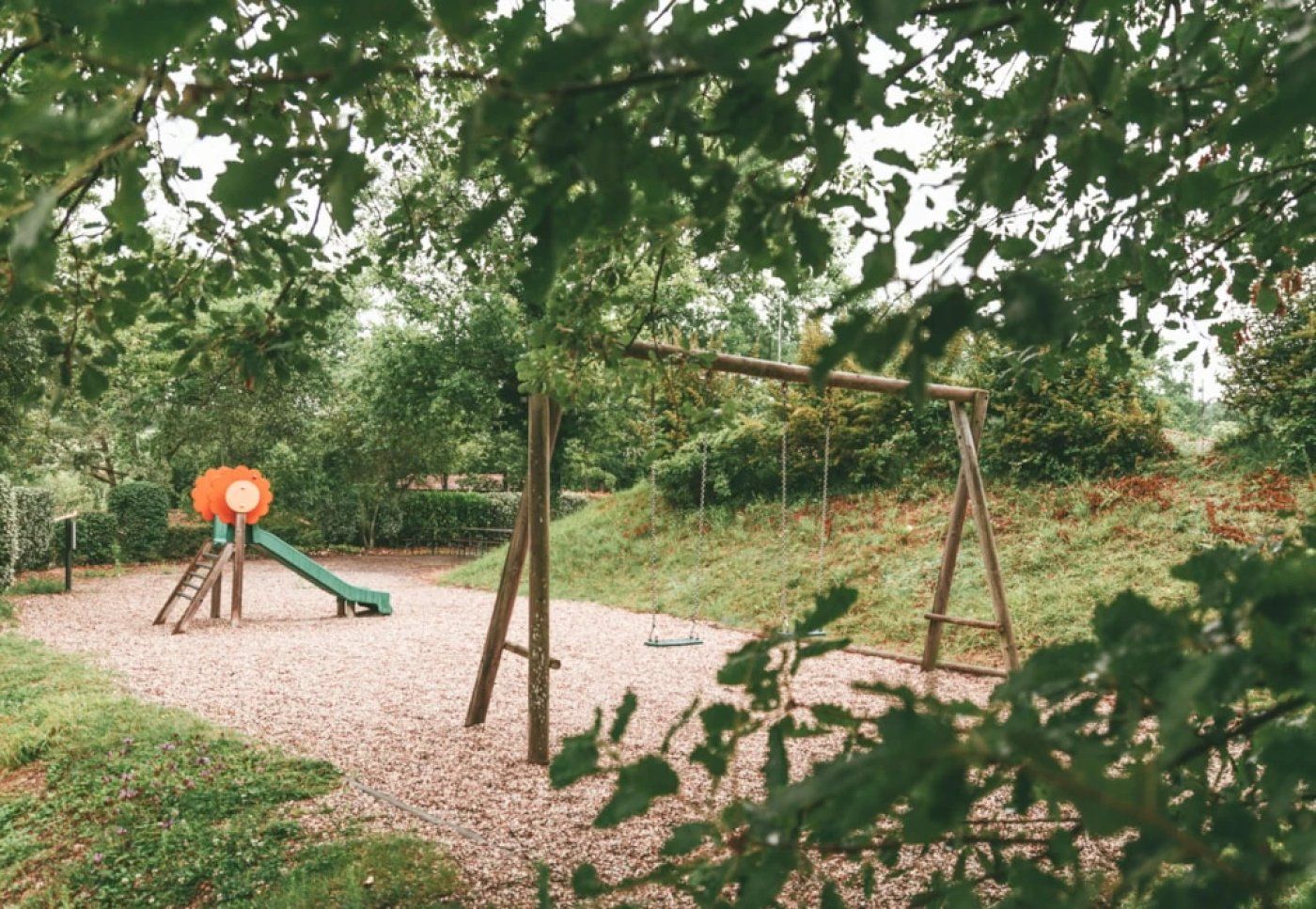 Een speeltuin met schommels en een glijbaan, omgeven door bomen en groen.