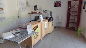 Un mostrador de recepción con una computadora, una silla y una pequeña mesa con documentos. Se ve una estantería con libros y una planta en maceta.
