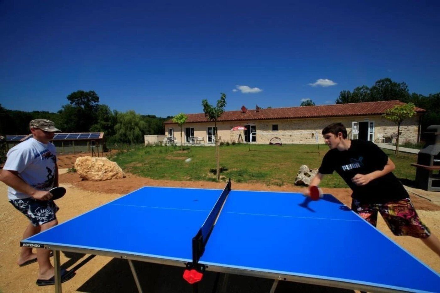 Twee personen spelen tafeltennis buiten, met een huis en zonnepanelen op de achtergrond.