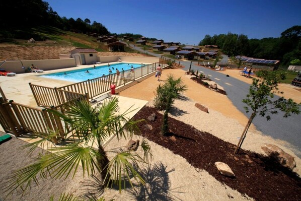 A swimming pool area with a sandy deck, palm trees, and a view of houses in the background.