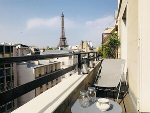 A balcony with a view of the Eiffel Tower, a white chair, and two glass jars on a small table. - Appartement vue Tour Eiffel Avec Terrasse (Paris)