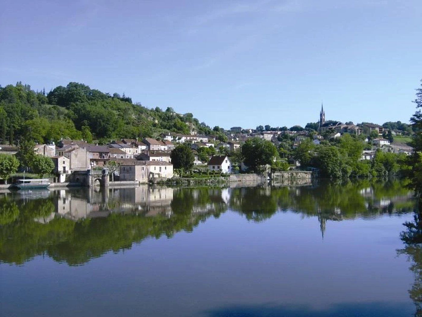 Une rivière paisible où se reflètent les bâtiments et la végétation, et où l’on aperçoit au loin le clocher d’une église.