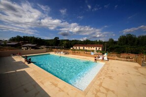 A rectangular swimming pool with a wooden deck surrounding it, a fenced area with lounge chairs, and a building in the background. - ZEcamping 3 étoiles piscine chauffée dog friendly cottage TV LV clim 35m2 (Cazals)