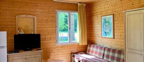 Une cabane en bois avec un téléviseur posé sur un meuble, un canapé, une table et deux fenêtres avec des rideaux.