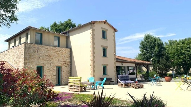 A two-story building with a stone facade and a tiled roof. There are outdoor seating areas with chairs and tables.