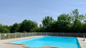 A rectangular swimming pool with a tiled deck, surrounded by a white fence and trees in the background.