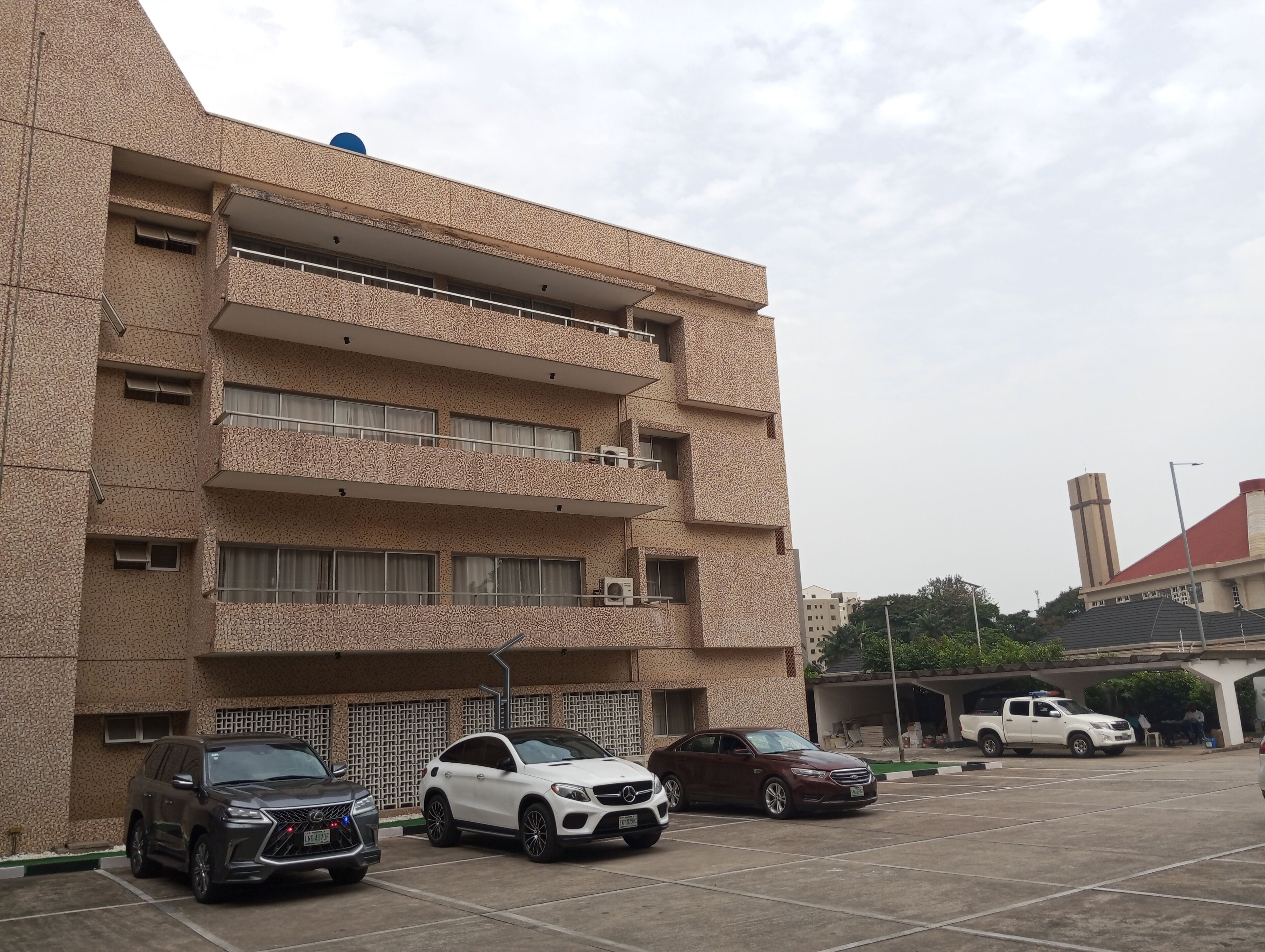 Un edificio de varios pisos con fachada texturizada, varios balcones y un estacionamiento con diversos vehículos.
