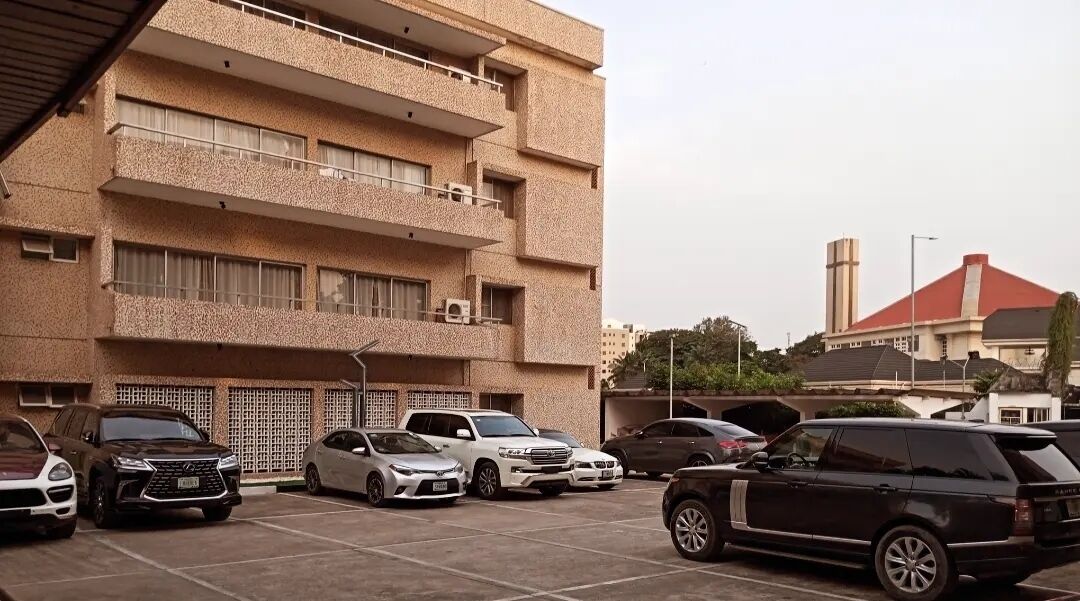 Un estacionamiento con varios coches, un edificio de varios pisos con balcones y una estructura de techo rojo al fondo.