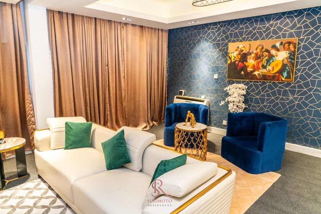 A modern living room with a white sofa, blue armchairs, a gold side table, and a large painting on the wall.