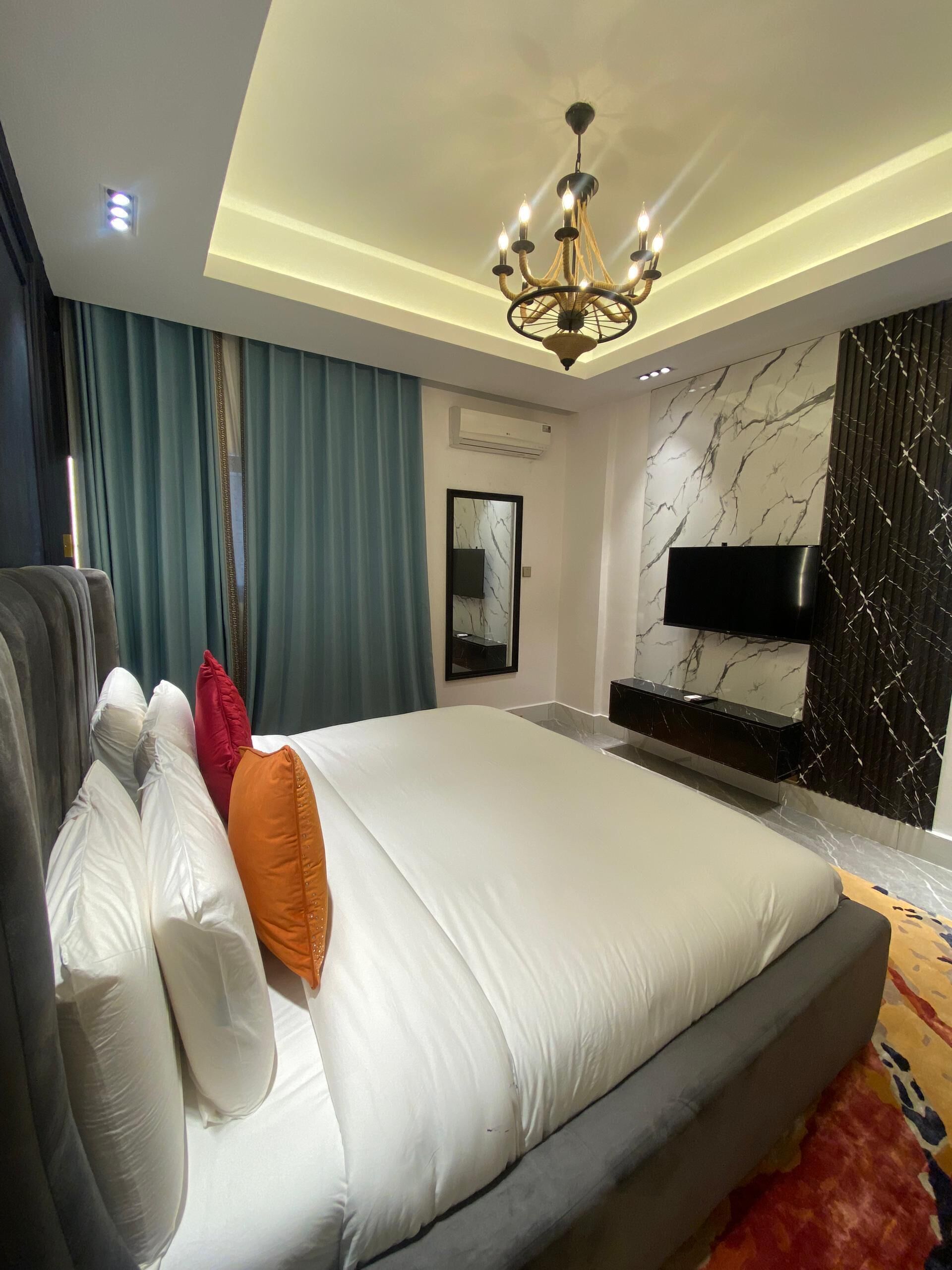 A modern bedroom with a large bed, a chandelier, a flat-screen TV, and a marble-patterned wall.