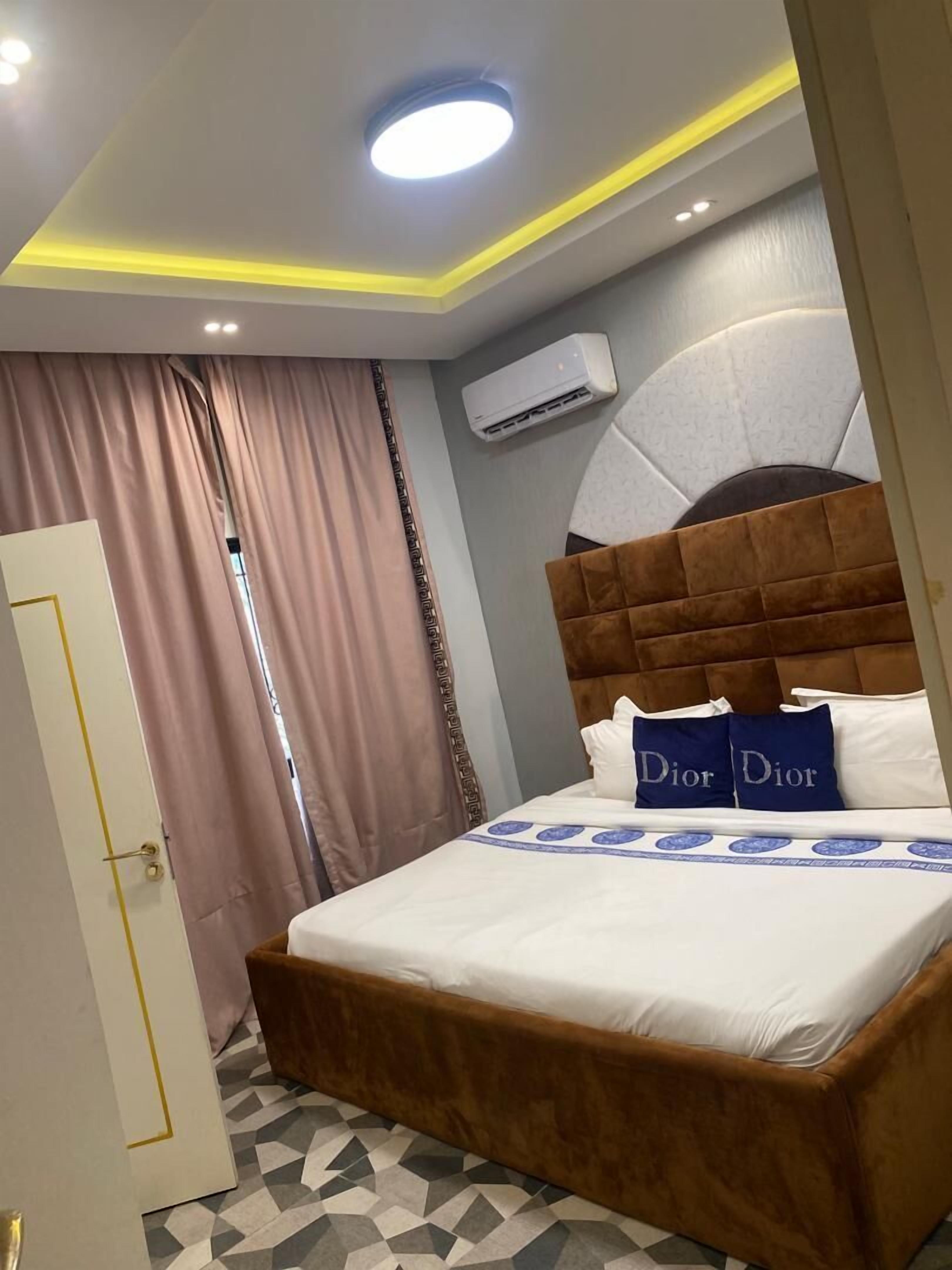 A hotel room with a large bed, a headboard, a ceiling light, an air conditioning unit, and a patterned floor.