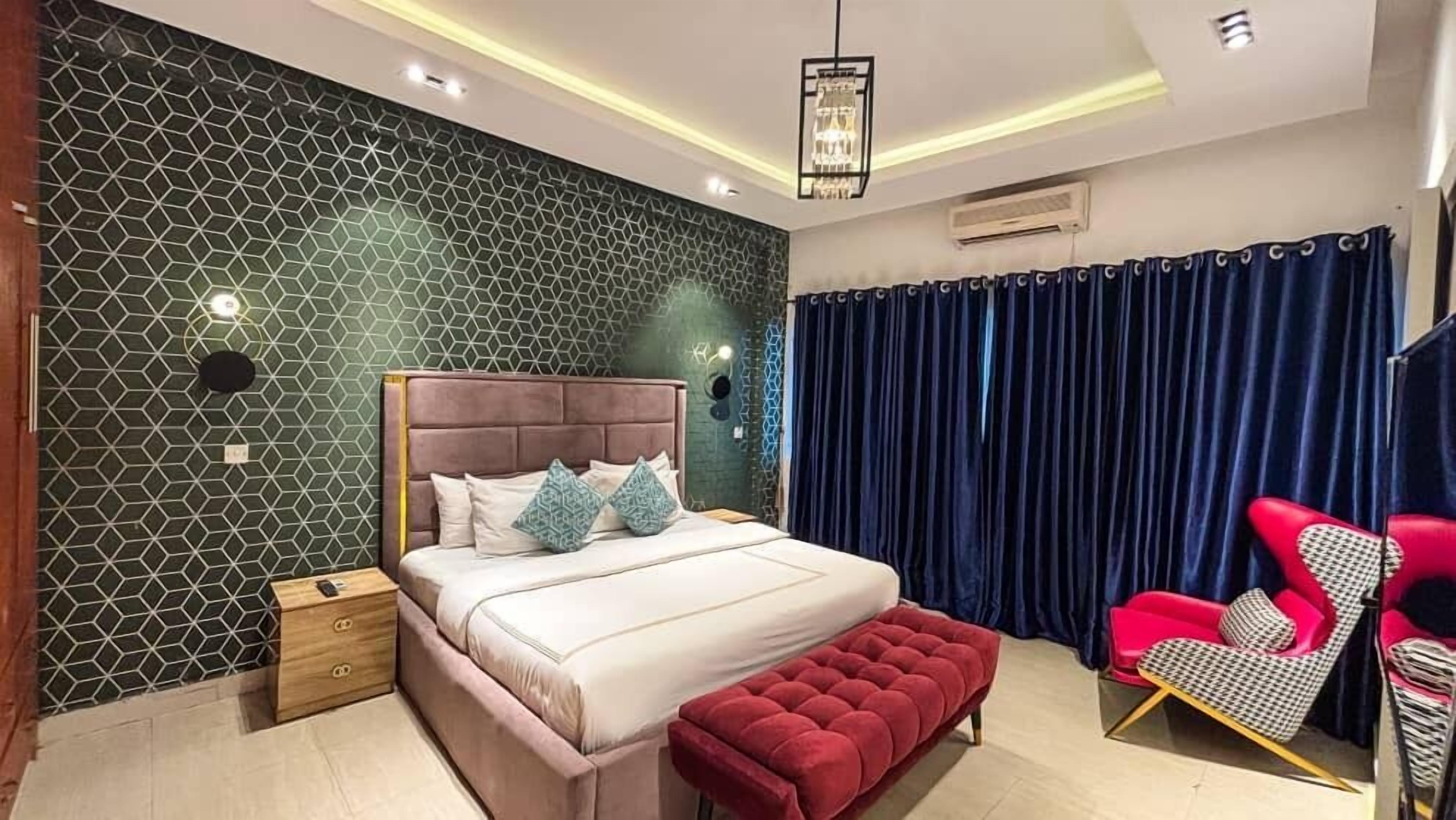 A modern bedroom with a geometric-patterned wall, a tufted red bench, a bed with white bedding, and a red and black patterned chair.