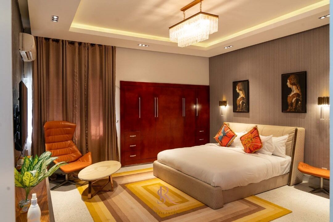 A modern hotel room with a large bed, a sitting area with a chair, a desk, and a wardrobe.