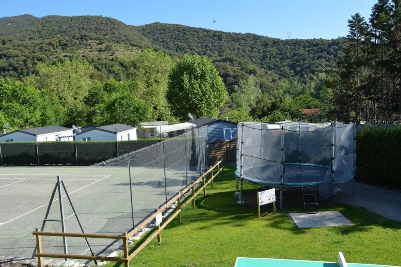 A tennis court with a net, surrounded by a fence, and a trampoline covered with a protective net.