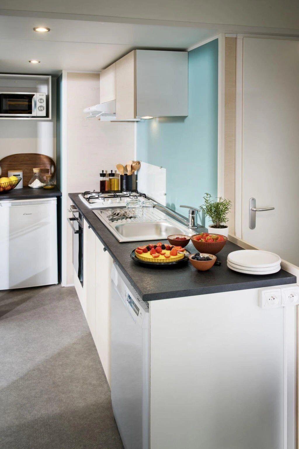 Una cocina compacta con muebles blancos, encimera negra y horno integrado. Se observan varios utensilios de cocina y un frutero en la encimera.