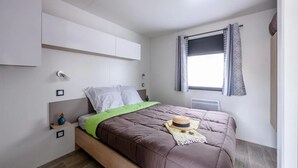 A bedroom with a bed, a window with curtains, a radiator, and a wall-mounted shelf.