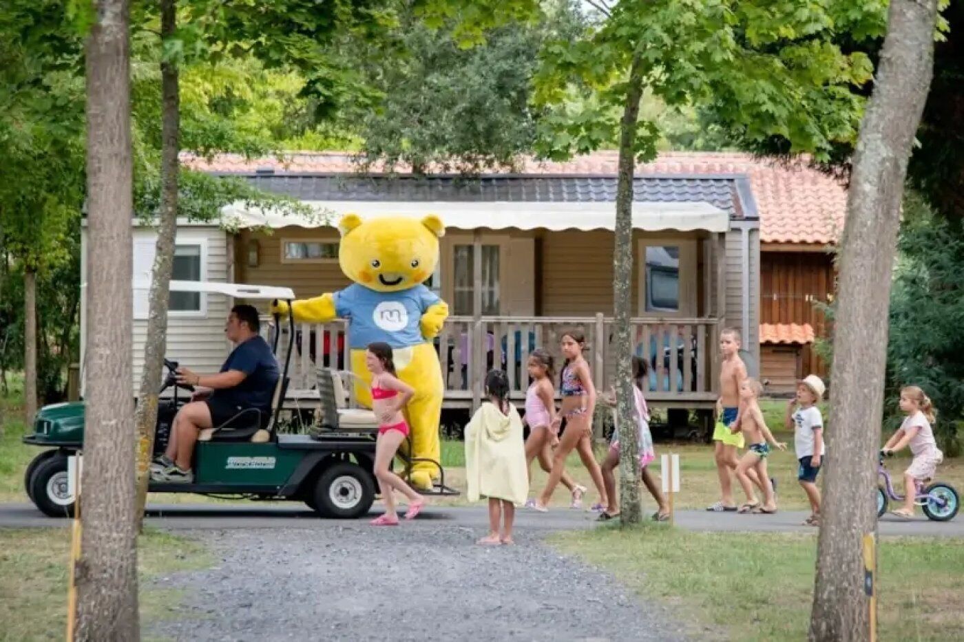 A person in a bear costume on a golf cart, surrounded by children and adults in a park-like setting.
