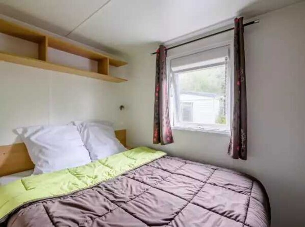 A single bed with a green and brown quilt, a wooden headboard, and a window with curtains.