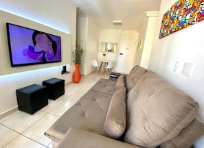 A living room with a large flat-screen TV, a beige sofa, a potted plant, and a dining area in the background.