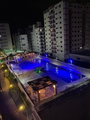 A nighttime view of a hotel with a swimming pool, lounge chairs, and a small garden area.
