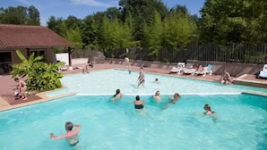 Ein Schwimmbeckenbereich mit badenden und spielenden Personen, umgeben von Bäumen und einem eingezäunten Bereich mit Liegestühlen.