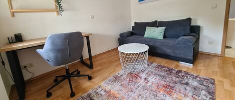 A modern living room with a grey office chair, a grey sofa with cushions, a round white side table, and a patterned rug.
