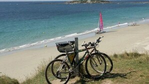 Un vélo garé sur une plage de sable, avec vue sur l’océan et un voilier à l’horizon.