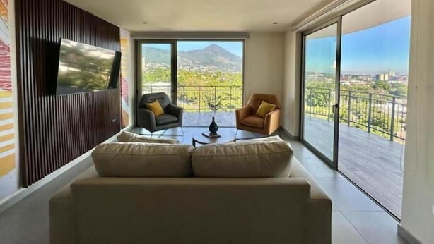 Een moderne woonkamer met een bank, twee fauteuils, een salontafel en een televisie bevestigd aan een houten wandpaneel. Grote schuifdeuren leiden naar een balkon met uitzicht op de bergen.
