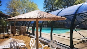 Un espace aménagé au bord de la piscine, avec un parasol en chaume, une terrasse en bois et des chaises en plastique blanches.
