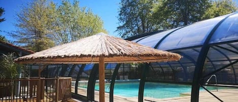 Zona de piscina con una sombrilla de paja, muelle de madera y sillas de plástico blanco.