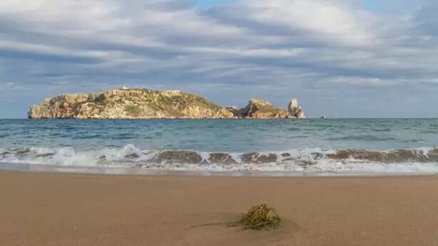 Una playa con un promontorio rocoso al fondo y olas acercándose a la orilla.