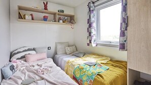 A small, well-lit bedroom with two beds, a window with purple curtains, and a shelf above the beds.