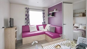 A modern, compact living space with a purple sofa, a small TV on a wooden stand, and a dining area with a table set for two.